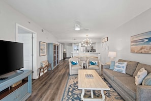 A living room with a television, sofa, coffee table, and a dining area with chairs and a chandelier.