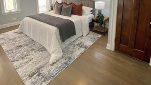A bedroom with a large bed, a wooden floor, a gray and white rug, a nightstand with a lamp, and a door.