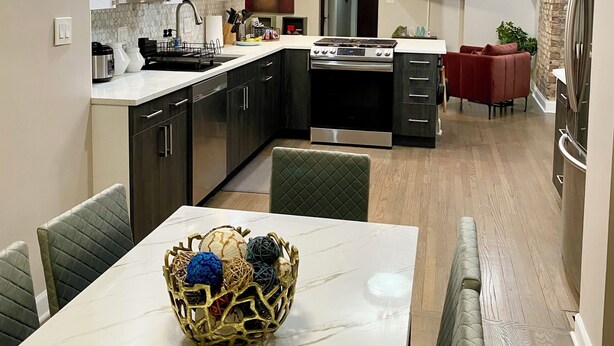 A modern kitchen with dark cabinets, a built-in microwave, and a stove. There is a dining area with a table and chairs, and a TV mounted on the wall.