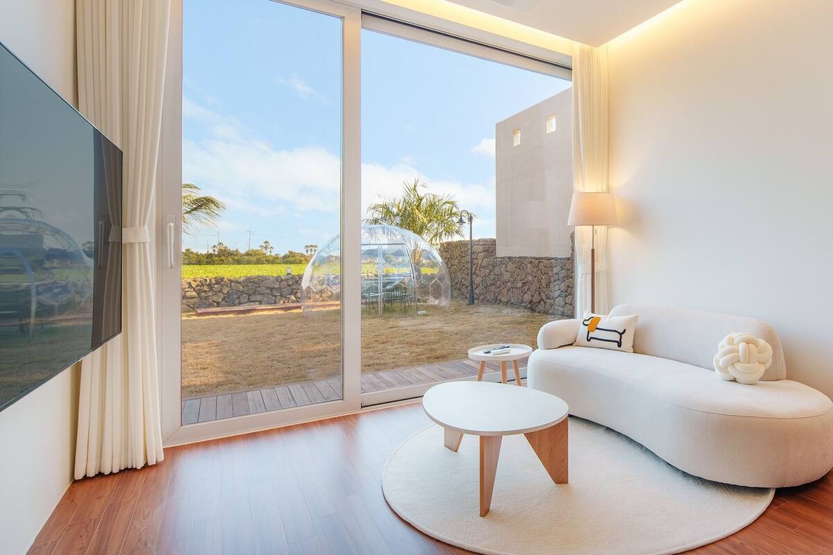 A modern living room with a glass door leading to a garden, a white sofa, a small round table, and a lamp.