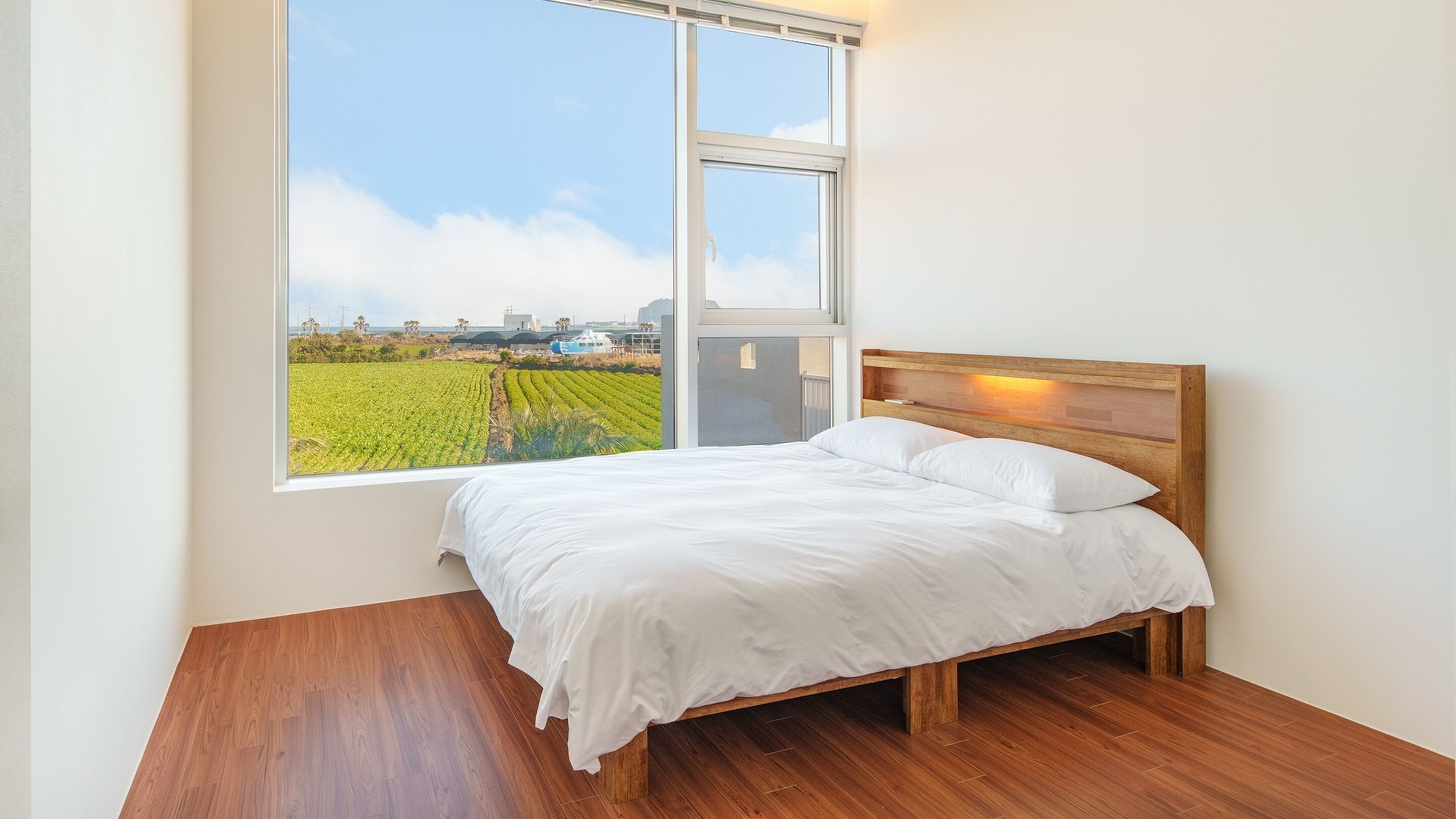 A bedroom with a large window, a wooden bed frame, and a view of a green field and buildings.