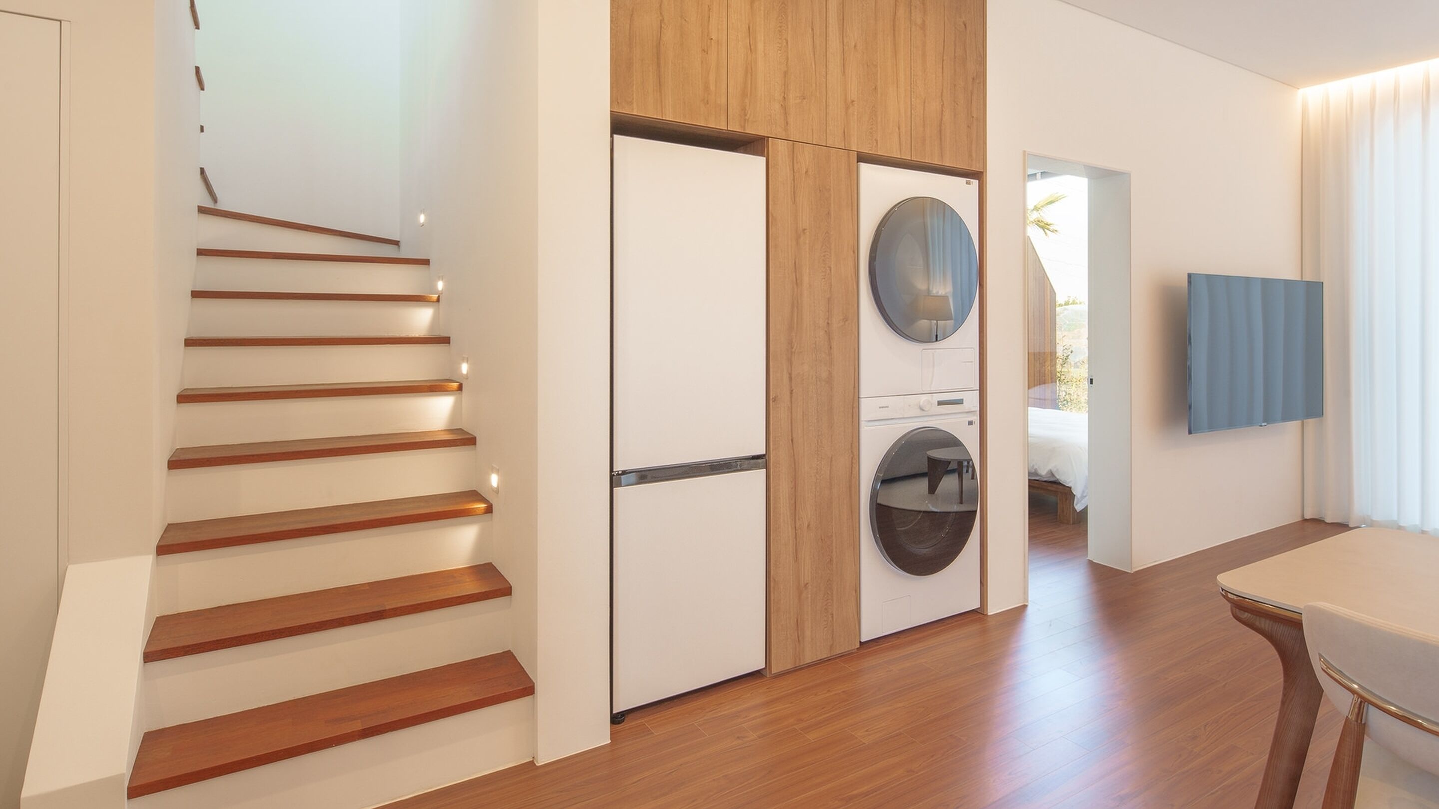 A modern interior with a wooden staircase, a laundry area, and a bedroom with a bed and a TV.