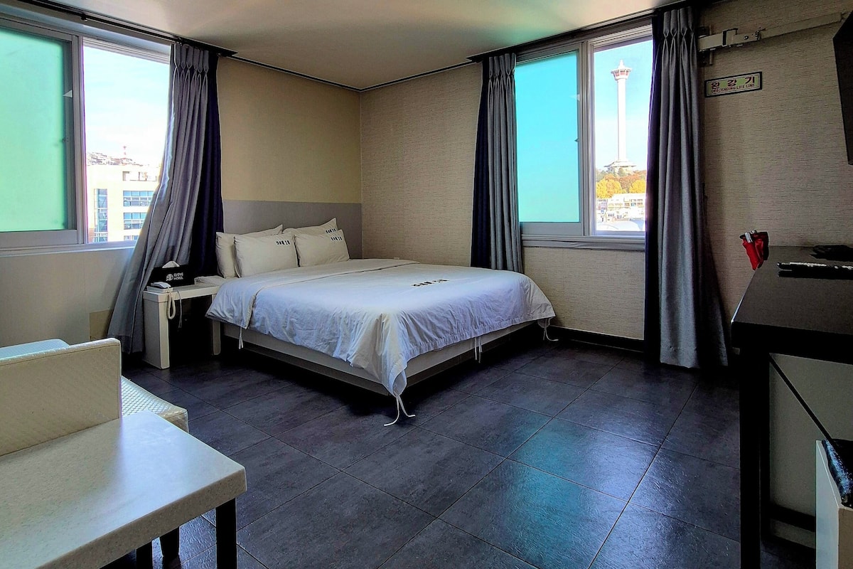 A hotel room with a bed, a desk, and a chair. There are curtains on the windows, and a sign on the wall.