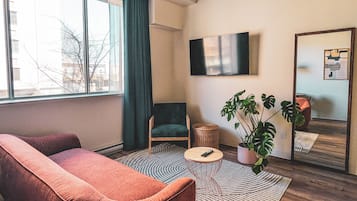Una sala de estar con un sofá, una silla, una mesa redonda, una planta en maceta, un espejo y un televisor en la pared.