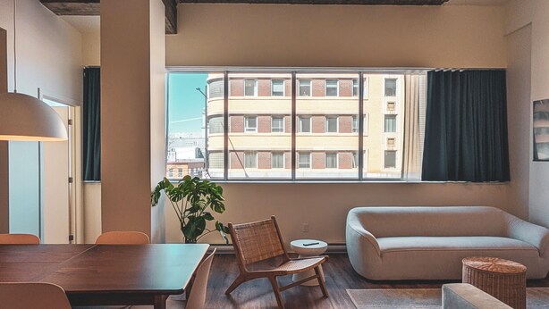 A modern living room with a wooden dining table, two chairs, a sofa, and a large window with a view of a building. - Terminus Hôtel (Québec)