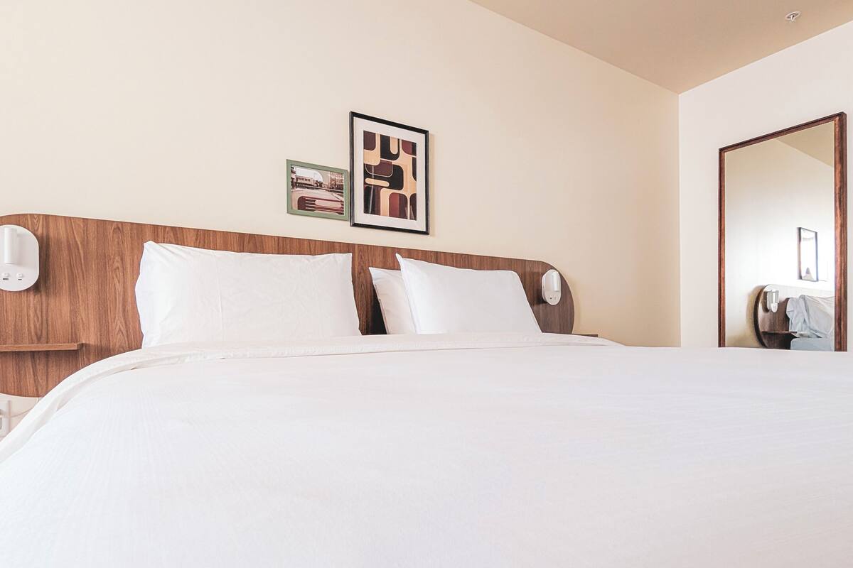 Quarto de hotel com cabeceira de madeira, uma cama grande com roupa de cama branca, duas fotografias emolduradas na parede e um espelho no lado direito.