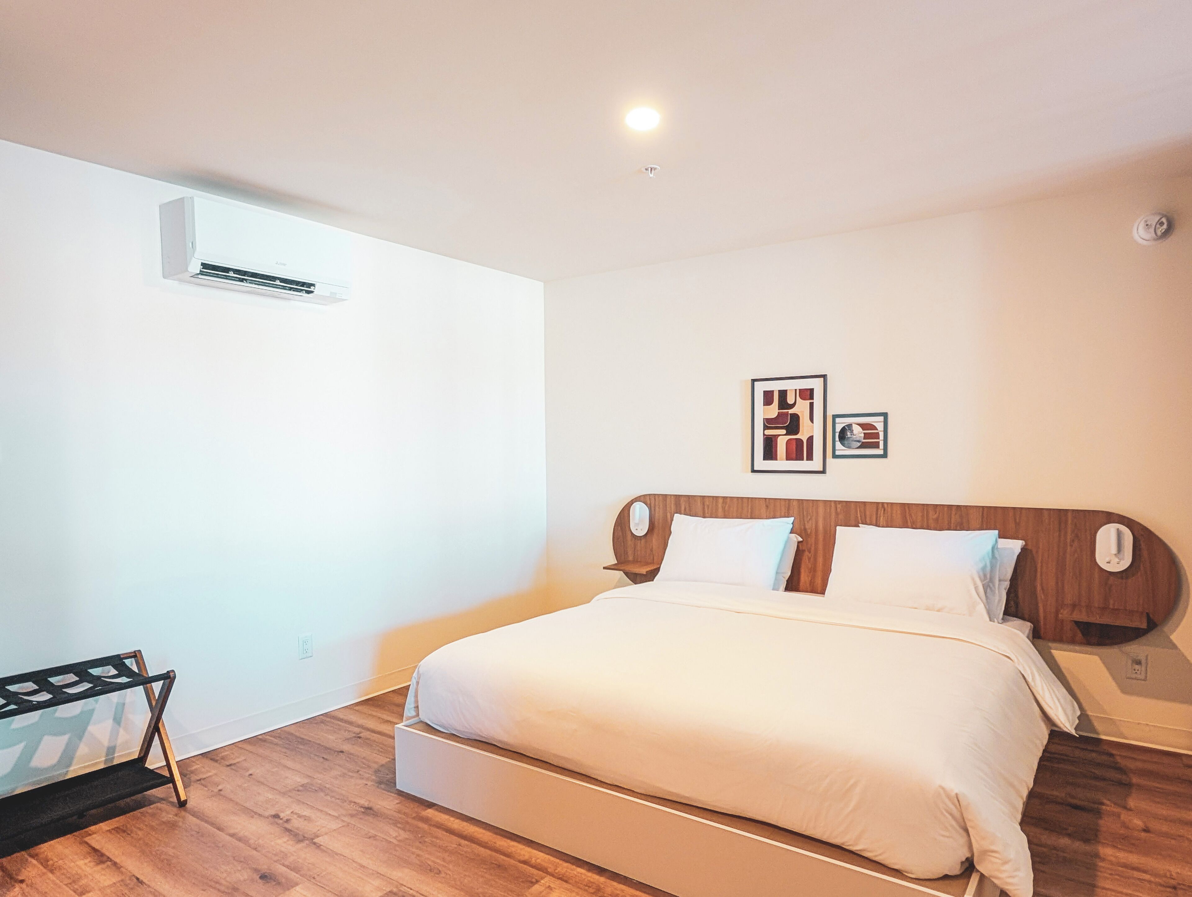 Un dormitorio con cabecera de madera, cama con ropa de cama blanca, una mesita de noche pequeña y un aire acondicionado instalado en la pared.