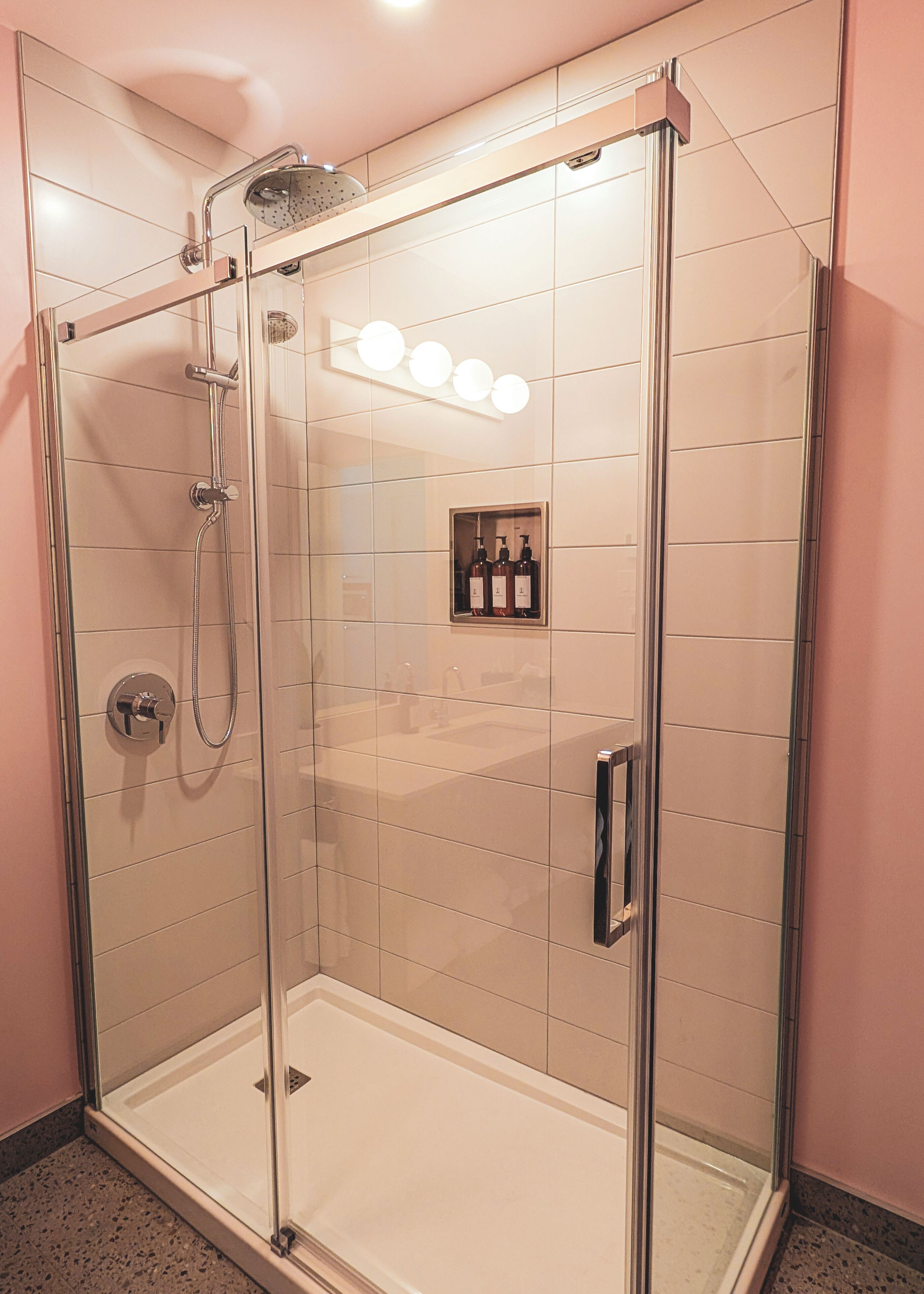 A glass-enclosed shower stall with a handheld showerhead, a control panel, and a shelf with toiletries.