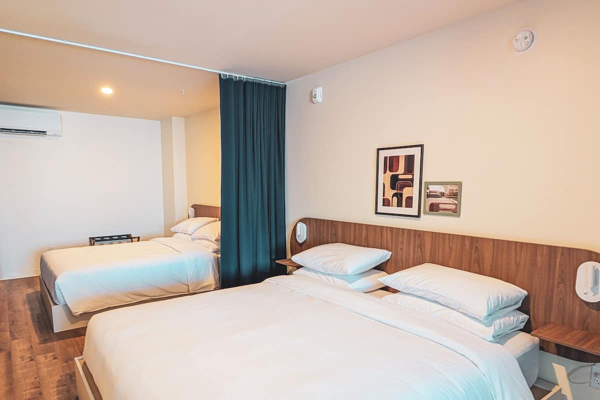 Quarto de hotel com duas camas de solteiro, cabeceira de madeira, cortina escura e ar condicionado fixado na parede.