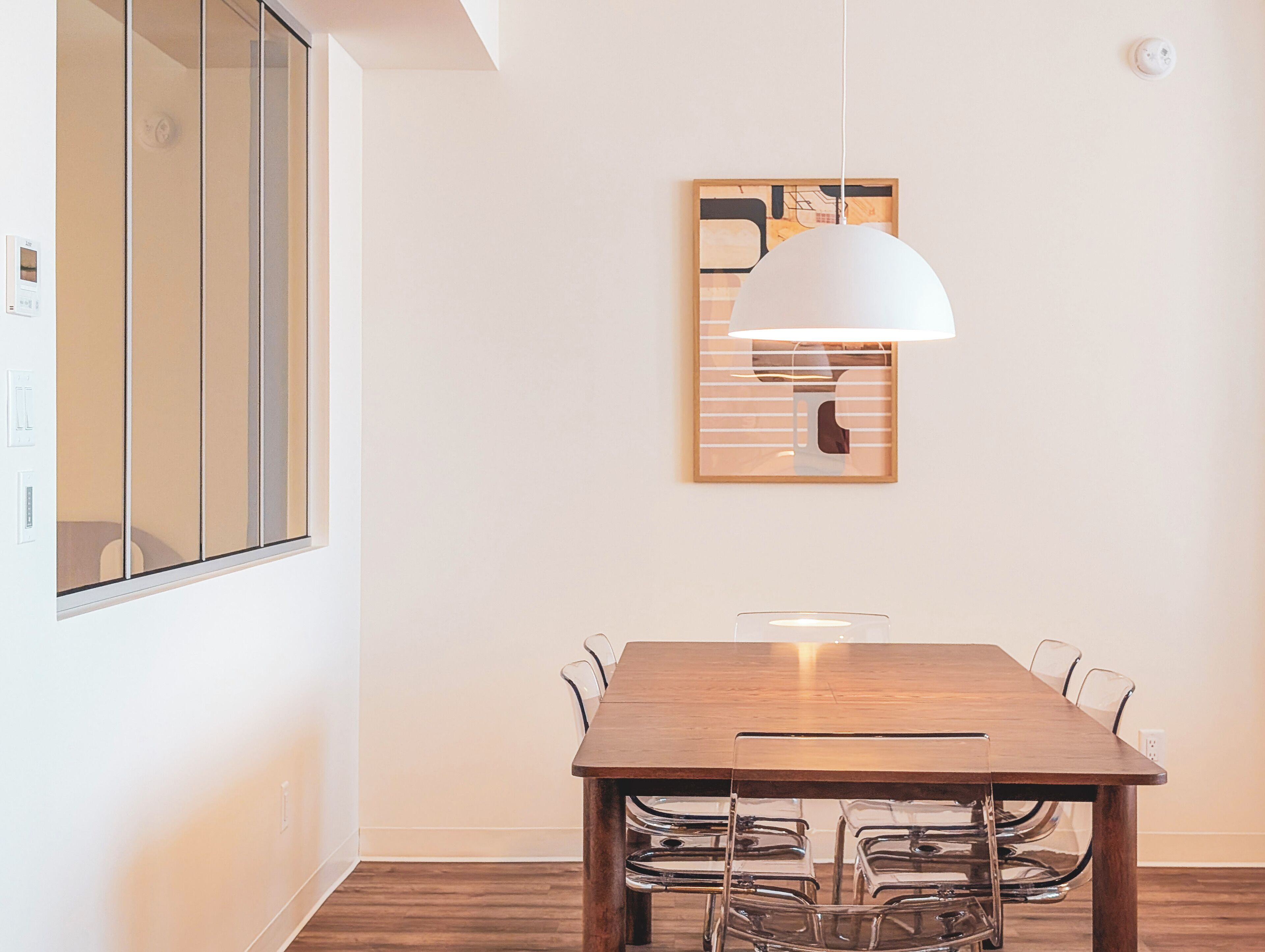 Un coin repas avec une table en bois et des chaises transparentes, un luminaire suspendu blanc et une œuvre d’art abstraite encadrée accrochée au mur.