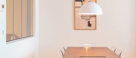 Un coin repas avec une table en bois et des chaises transparentes, un luminaire suspendu blanc et une œuvre d’art abstraite encadrée accrochée au mur.