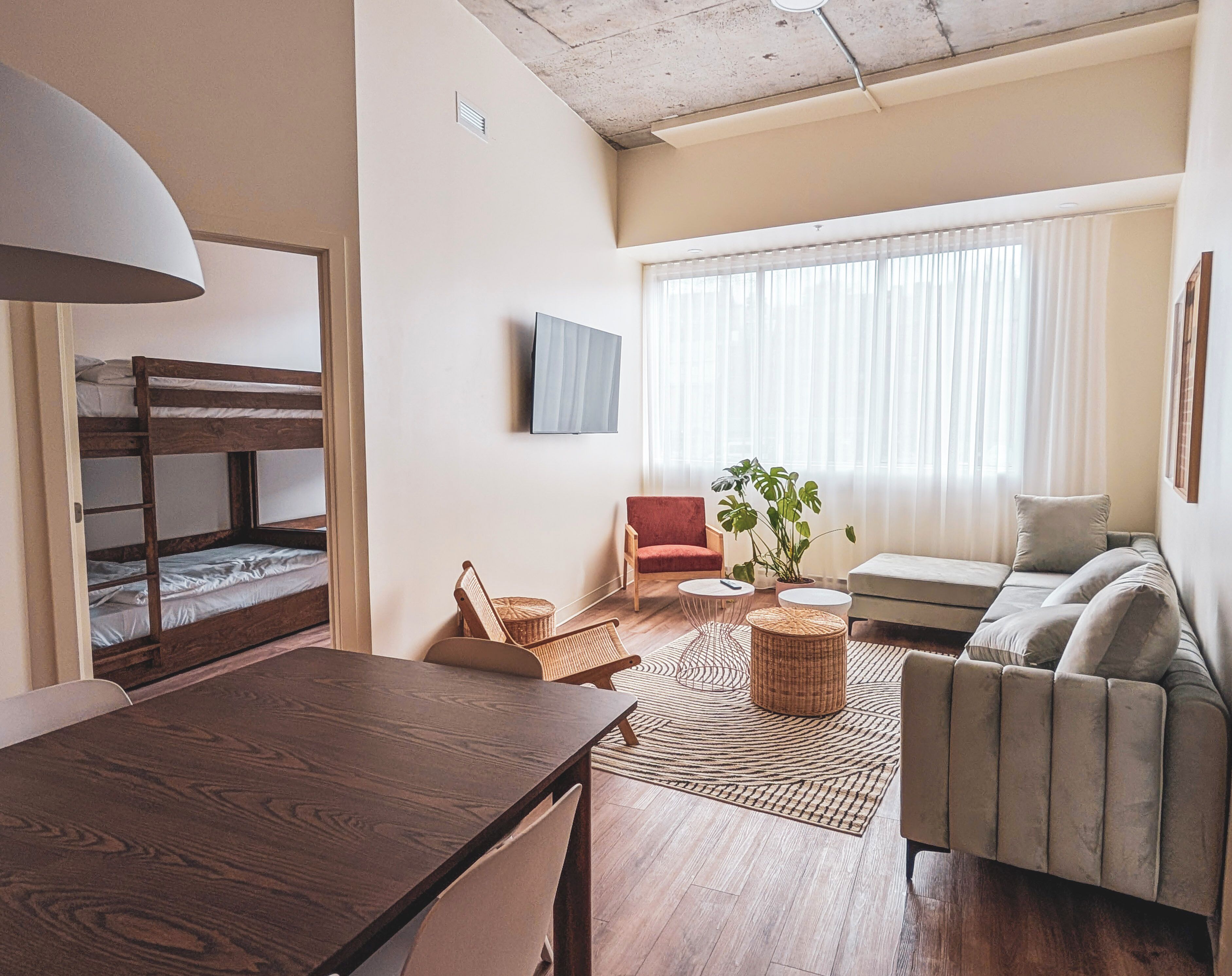 Una habitación con literas, una mesa de comedor, un sofá, una silla roja, un televisor y un ventanal con cortinas.