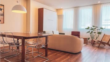Una sala moderna con una mesa de comedor de madera, sillas traslúcidas, un sofá y un ventanal con cortinas translúcidas.