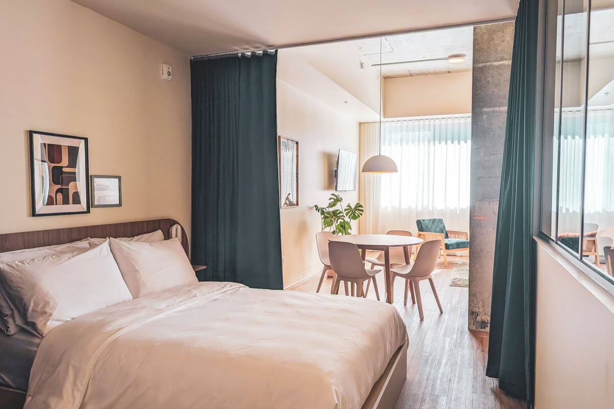 Quarto de hotel com cama, mesa de jantar com cadeiras, planta em vaso e vista para a varanda.