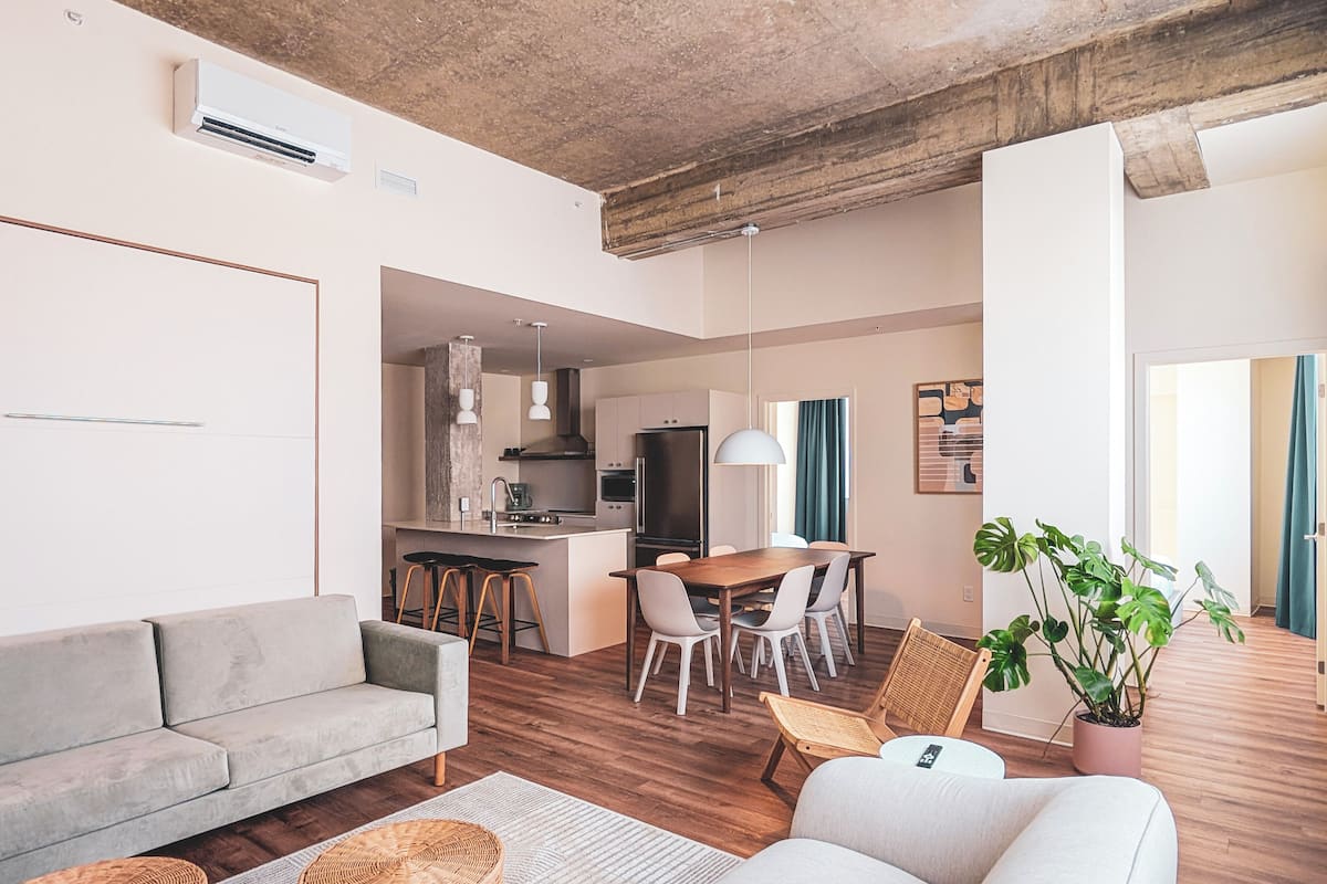 Uma sala moderna com sofá, área de jantar com cadeiras, cozinha com fogão e geladeira, e uma janela grande com cortinas.
