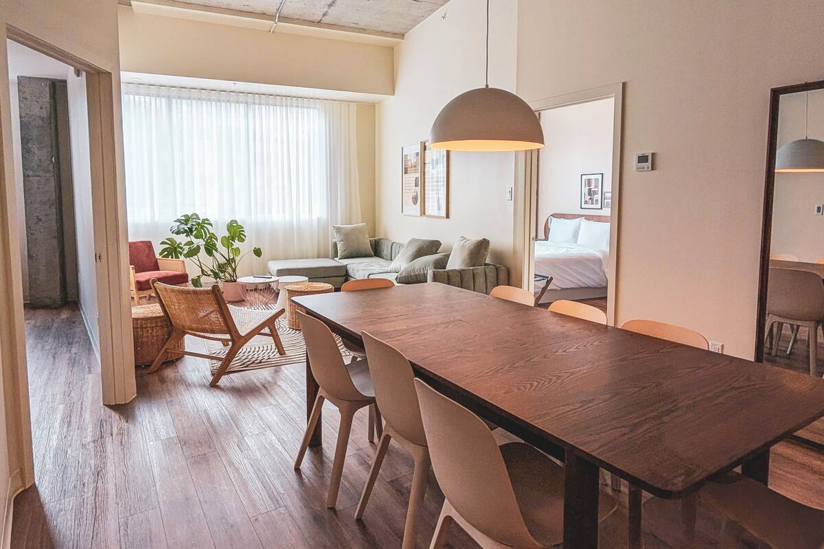 Uma sala de estar moderna com uma mesa de jantar de madeira, assentos acolchoados e uma janela grande com cortinas leves.