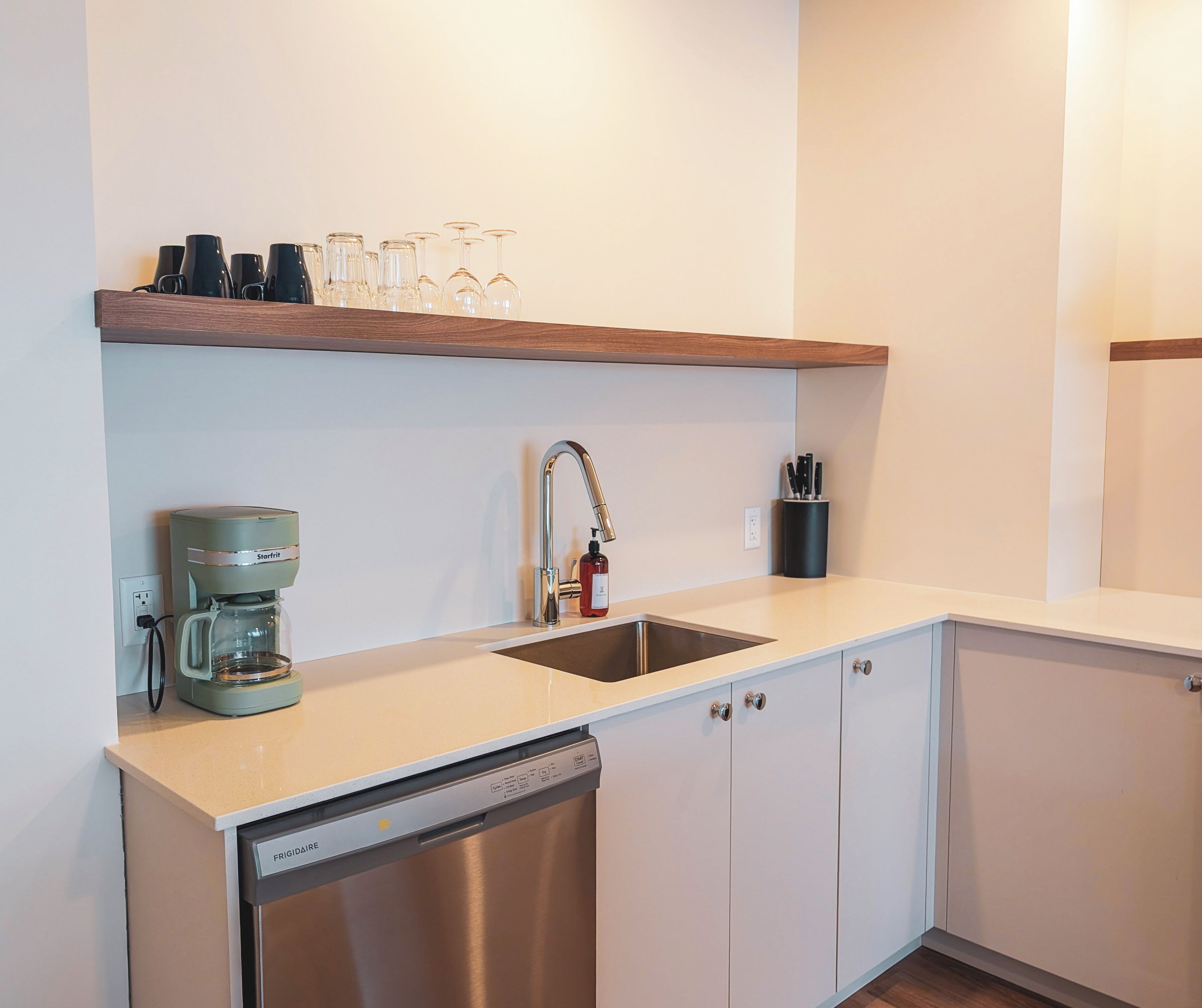 A modern kitchen with a built-in dishwasher, a coffee maker, a stainless steel sink, and a shelf with glassware.