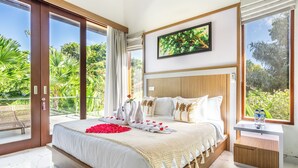Ein geräumiges Schlafzimmer mit einem großen Bett, einem hölzernen Kopfteil und Blick auf üppiges Grün durch Glastüren.