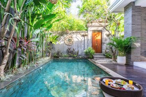 A clear swimming pool surrounded by lush greenery, a wooden door, and a dining area with a tray of food. - Villa Sun Lila (by JB Villas) (Seminyak)