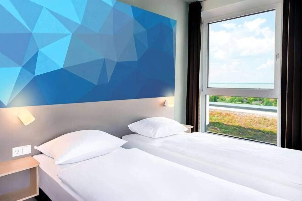 A hotel room with a geometric wall design, a bed with white bedding, a nightstand, and a window with a view of greenery.