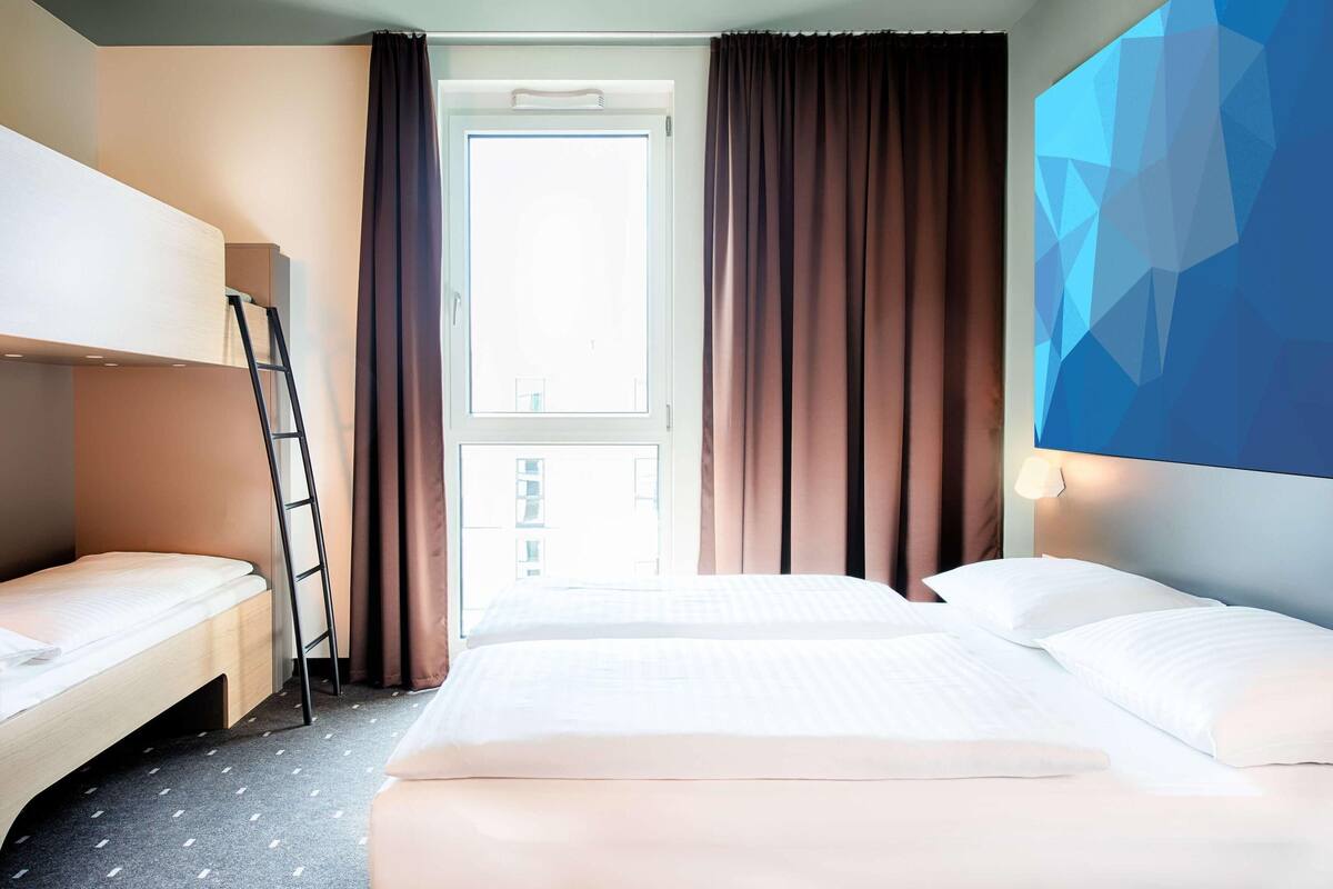 A hotel room with a bunk bed, a desk, and a large window with curtains.