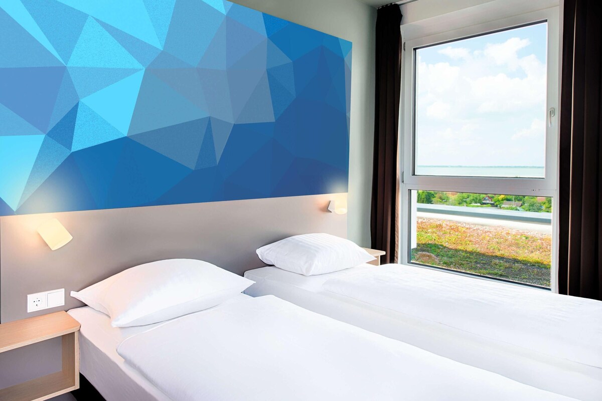 Habitación de hotel con cama, mesita de noche, lámpara de pared y ventana con vista a follaje.