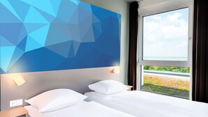 Habitación de hotel con cama, mesita de noche, lámpara de pared y ventana con vista a follaje.