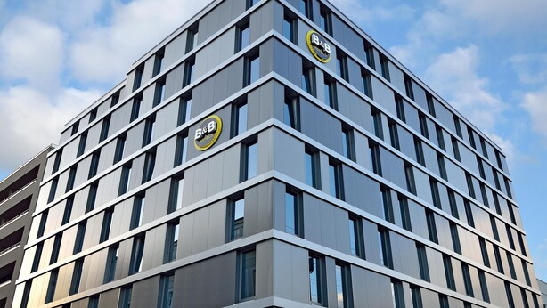 Un hôtel moderne, avec une façade ponctuée de nombreuses fenêtres et deux enseignes bien visibles indiquant qu’il s’agit d’un établissement proposant à la fois hébergement et petit-déjeuner.