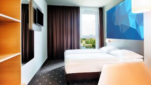 Une chambre d’hôtel avec un lit, un téléviseur fixé au mur, une fenêtre avec des rideaux et une vue sur la verdure à l’extérieur.