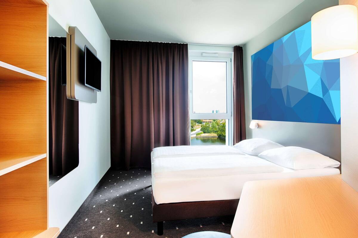 A hotel room with a bed, a TV mounted on the wall, a window with curtains, and a view of greenery outside.