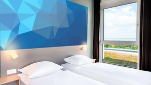 Habitación de hotel con cama, mesita de noche, lámpara de pared y ventana con vistas a zonas verdes.