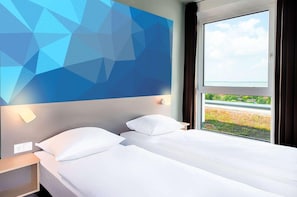 Habitación de hotel con cama, mesita de noche, lámpara de pared y ventana con vistas a zonas verdes.