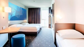 Une chambre d’hôtel avec deux lits, un bureau, un pouf bleu et un motif géométrique sur le mur.