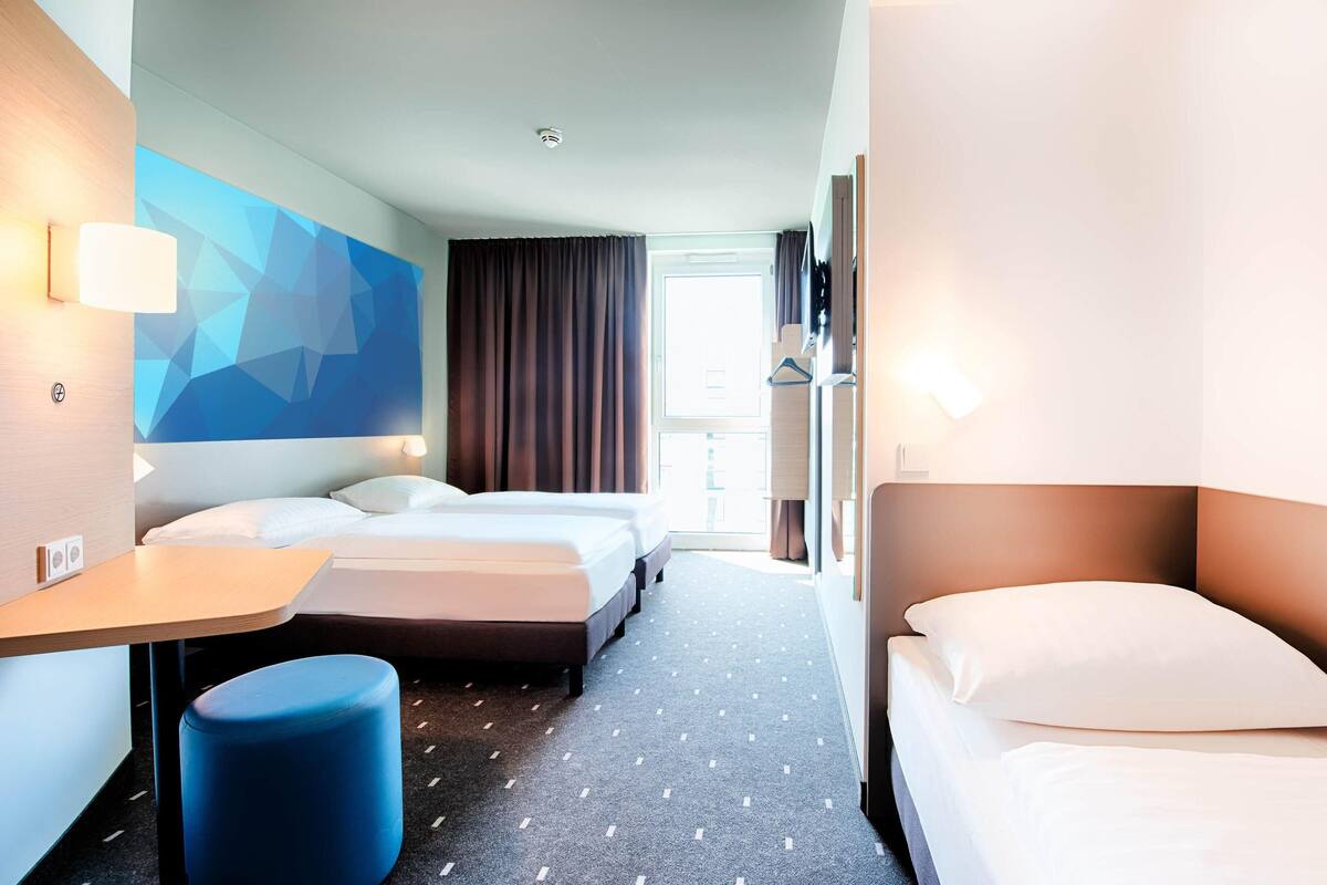 A hotel room with two beds, a desk, a blue ottoman, and a geometric wall design.