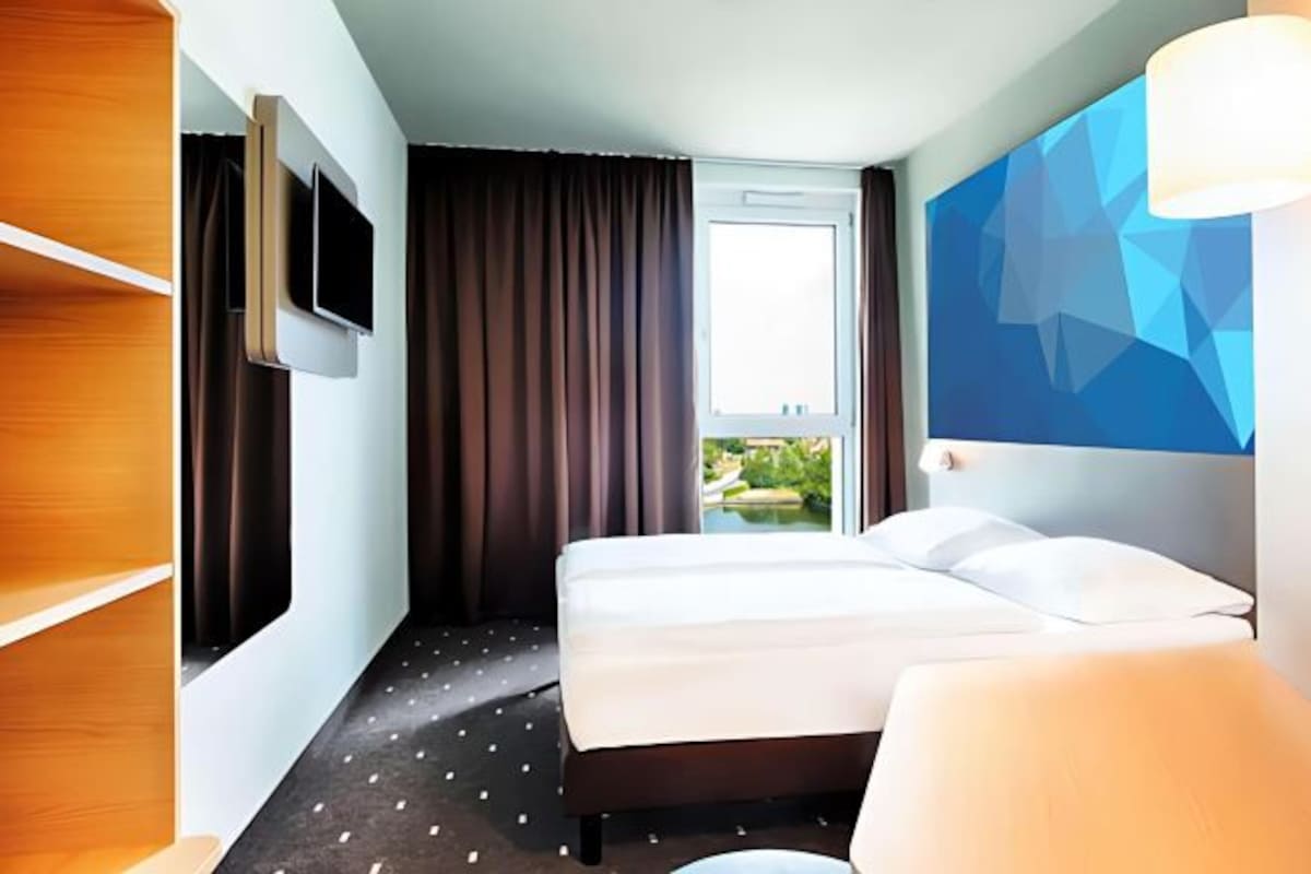 Habitación de hotel con cama, televisor montado en la pared, ventana con cortinas y vista a zonas verdes.