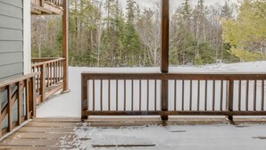 Une terrasse en bois dotée d’une rambarde, qui mène à un porche abrité offrant une vue sur une zone boisée.