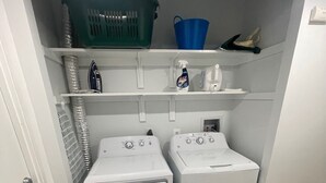 A laundry room with two white front-loading washing machines, a green and a blue laundry basket, and a shelf with cleaning supplies.