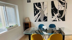 A dining area with a glass table and two yellow chairs, a window with a plant, and abstract wall art.