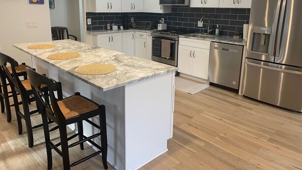 A modern kitchen with white cabinets, a central island with stools, stainless steel appliances, and a black tiled backsplash.