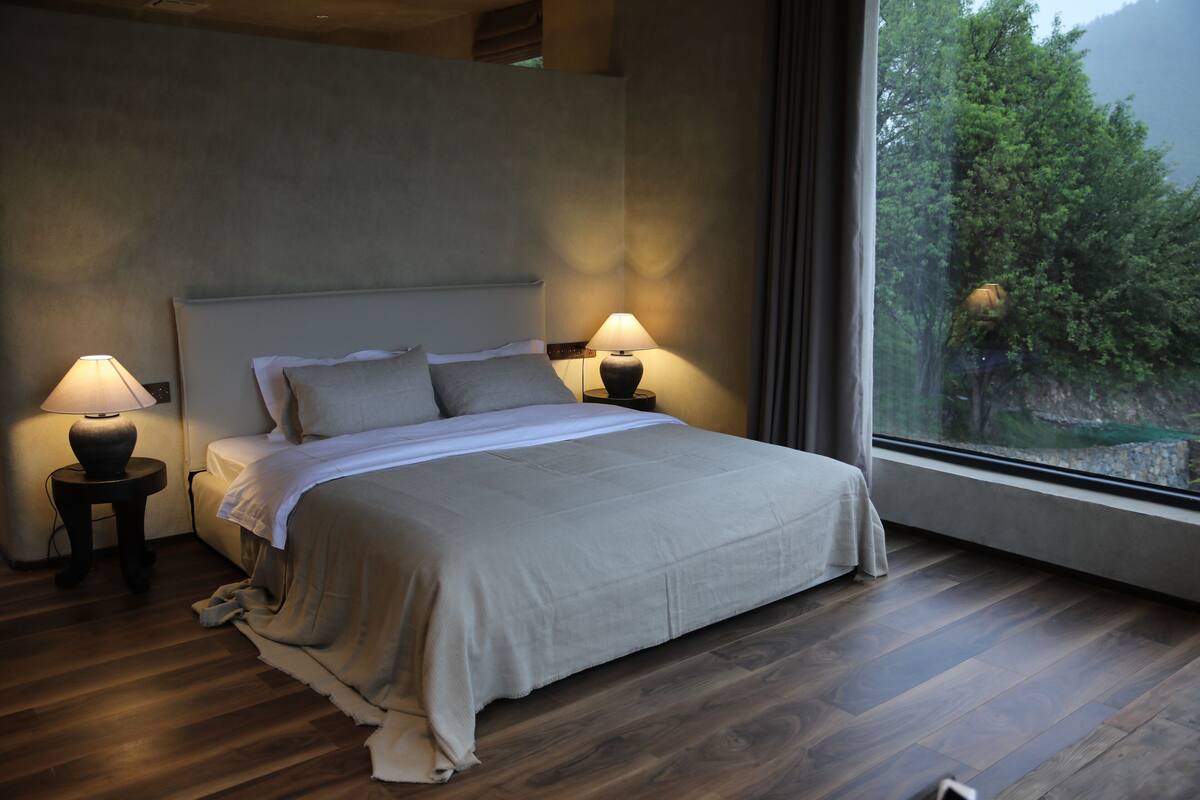Un dormitorio moderno con una cama grande, dos mesitas de noche con lámparas y vistas a zonas verdes a través de un ventanal amplio.