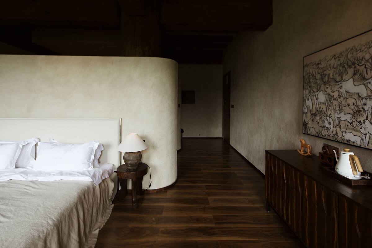 Habitación de hotel con cama, mesita de noche con lámpara, suelo de madera, pared curva y un cuadro en la pared.