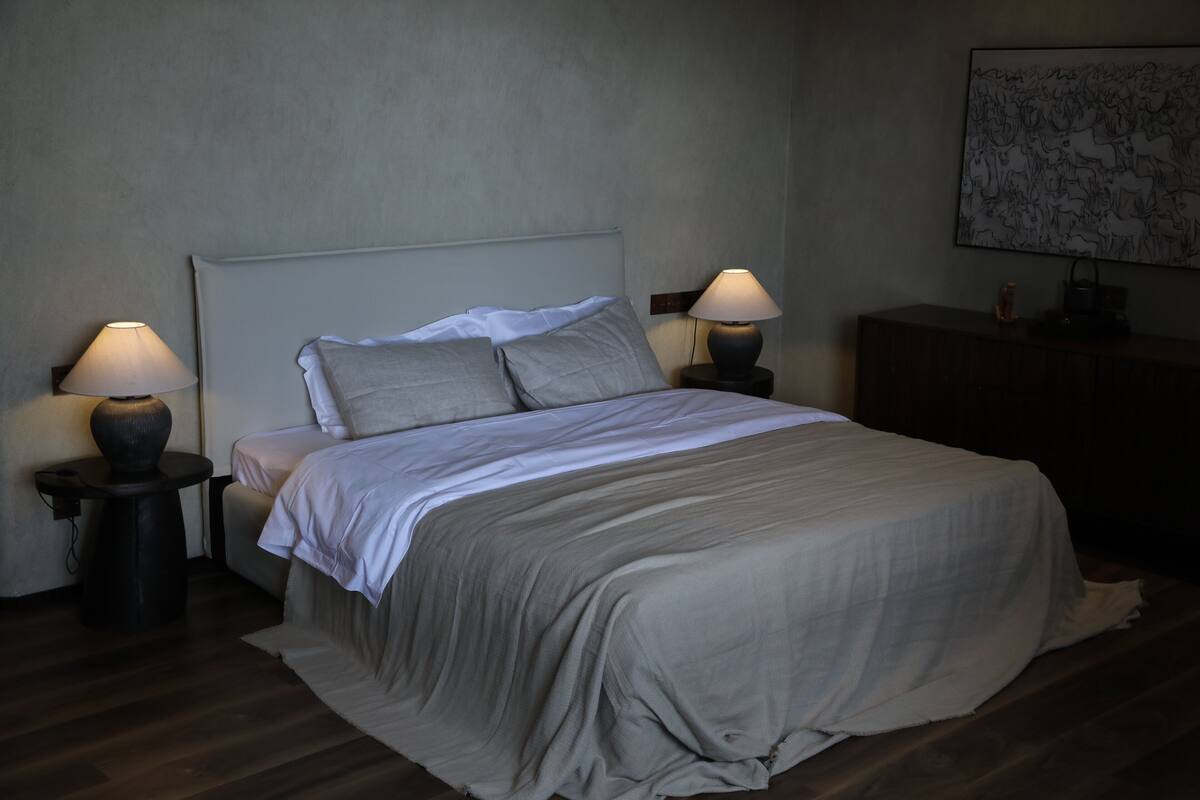 Una cama bien tendida con una colcha gris y almohadas, dos lámparas de noche, un tocador de madera y un cuadro en la pared.