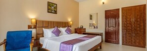 A hotel room with a bed, a nightstand, a blue chair, a wardrobe, and a mirror. - Sterling Brookstone Coorg (Madikeri)