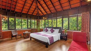 Un'ampia camera da letto con un letto grande, un'area salotto con una poltrona e un tavolo, e ampie finestre che offrono vista sul verde.