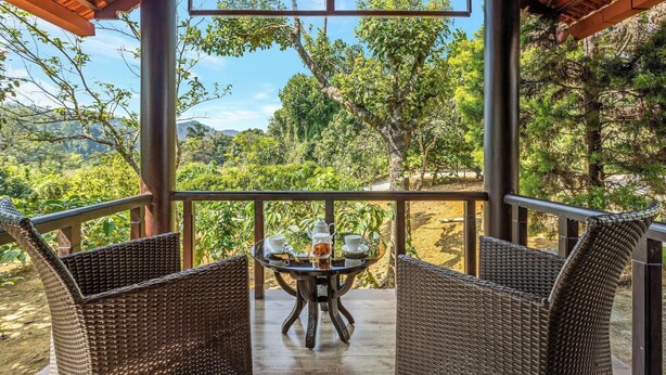 Un'area relax esterna coperta, con sedie in vimini e un tavolo, con vista su un giardino rigoglioso e dolci colline lontane.