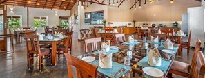 A spacious dining area with multiple tables and chairs, large windows offering a view of the outdoors, and a modern kitchen in the background. - Sterling Brookstone Coorg (Madikeri)