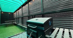 Una parrilla para barbacoa en un área exterior con piso de césped artificial verde, una mesa de picnic y persianas metálicas al fondo.