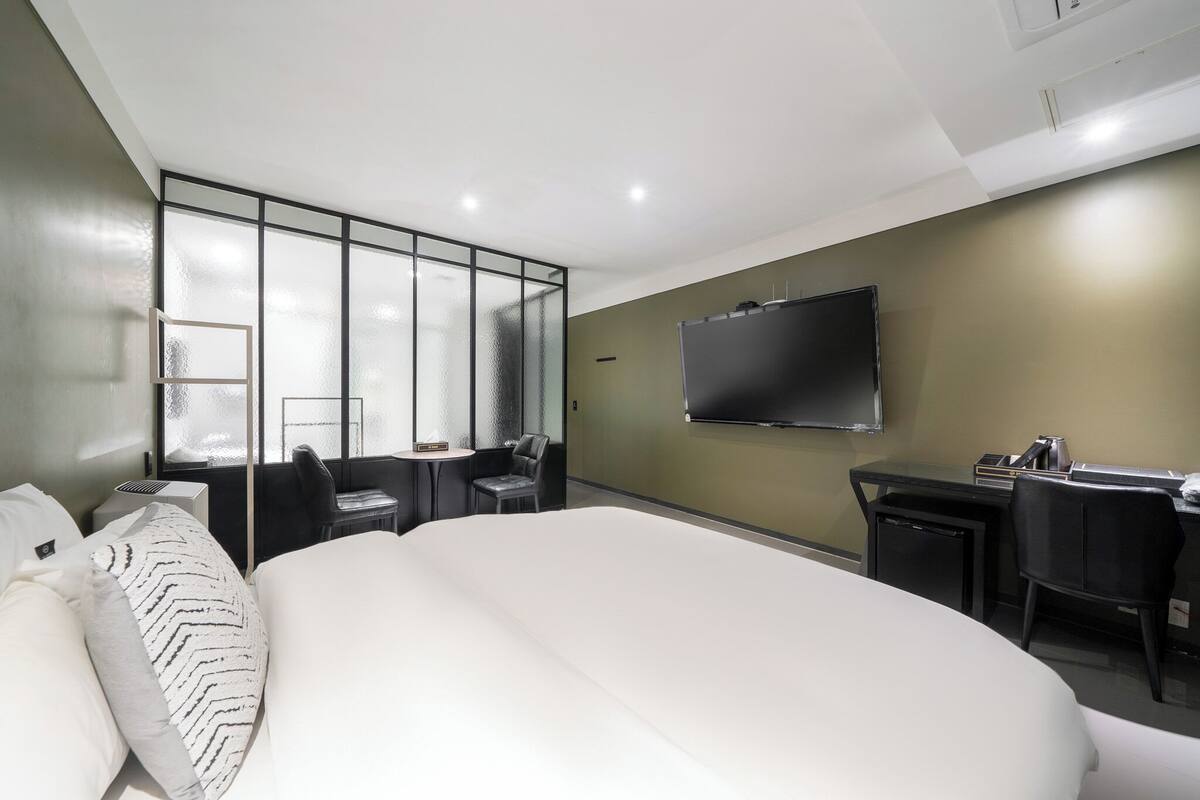 A modern hotel room with a large bed, a desk with a chair, a TV mounted on the wall, and a large window with a view.