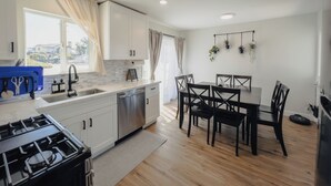 A modern kitchen with white cabinets, a stainless steel dishwasher, a gas stove, and a dining area with a wooden table and chairs. - Serene river view & beach walk, near Monterey Aquarium, Laguna Seca, Santa Cruz (Moss Landing)