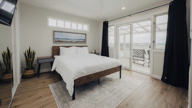 A bedroom with a large bed, a nightstand, a painting, and a view of the outdoors through a sliding door. - Serene river view & beach walk, near Monterey Aquarium, Laguna Seca, Santa Cruz (Moss Landing)